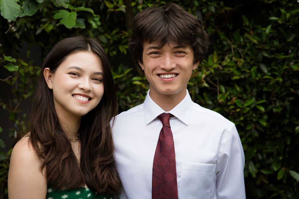 A young couple in formal attire smiles warmly in front of a green hedge backdrop.