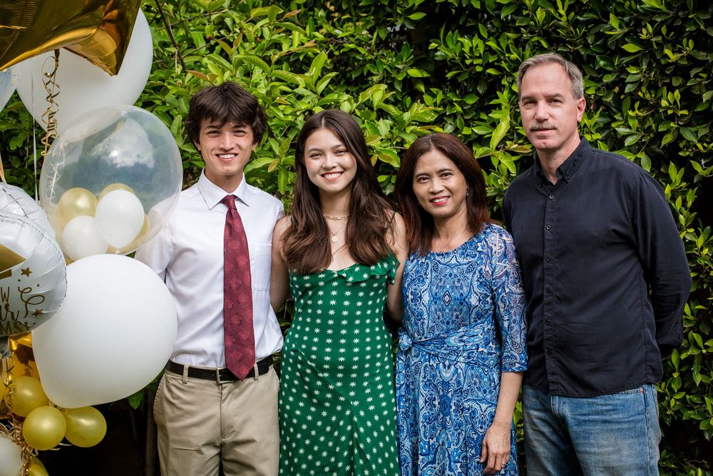 Group stands together at an outdoor event with green foliage in background and festive white and gold balloons.
