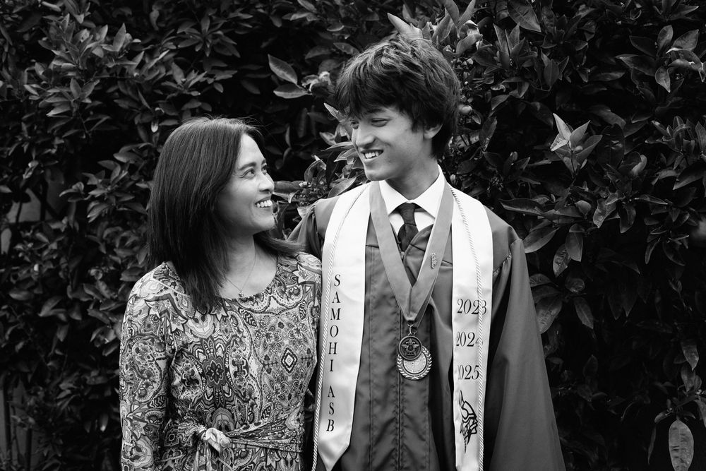 Black and white photo of a graduate in academic regalia and sash smiling with another person against a leafy background.