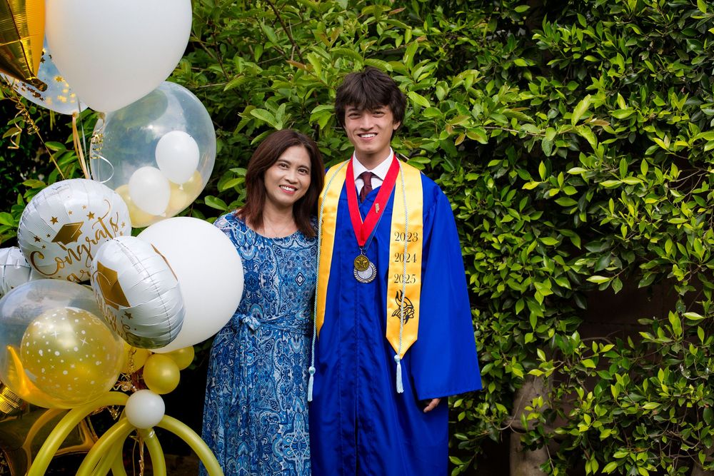 Graduate in royal blue cap and gown stands with balloons against green hedge at graduation celebration event.