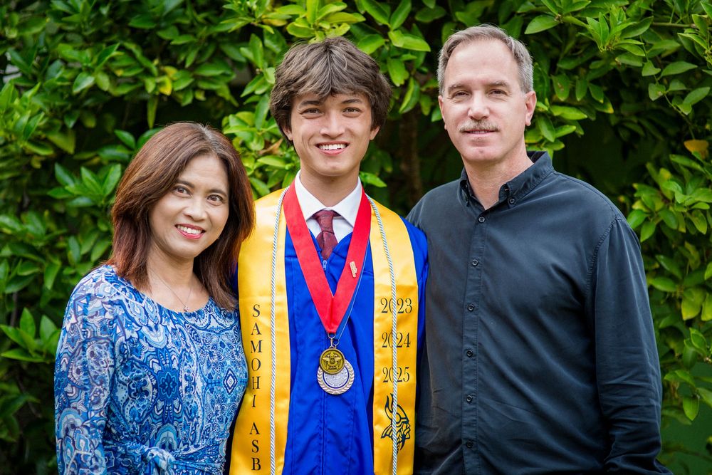 An outdoor family portrait shows three people smiling together against a backdrop of lush green foliage.