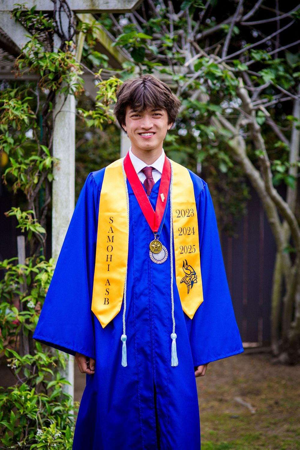 Graduate wearing blue academic regalia with gold stoles and a medal poses in front of garden foliage.