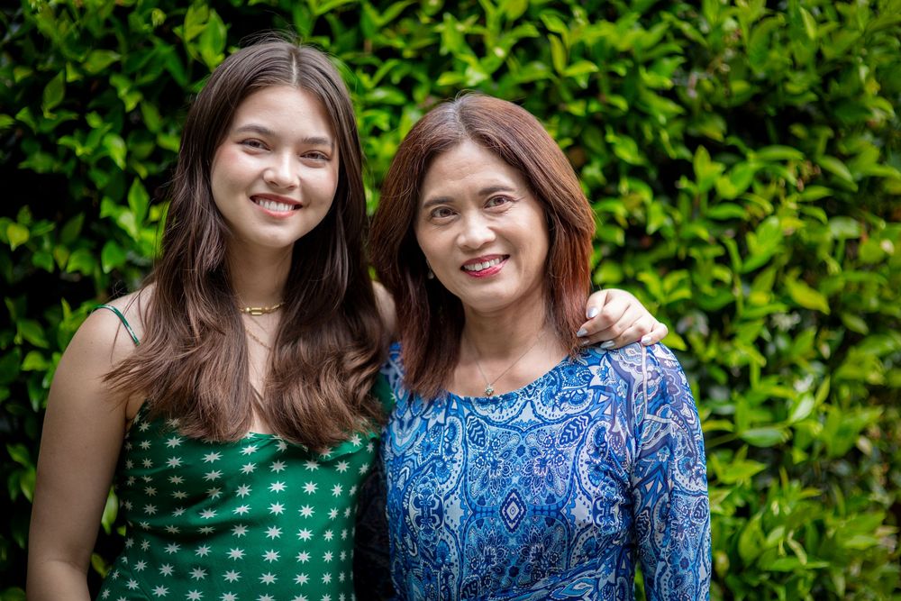 Two people wearing green polka dot dress and blue floral top pose together outdoors against lush green foliage.