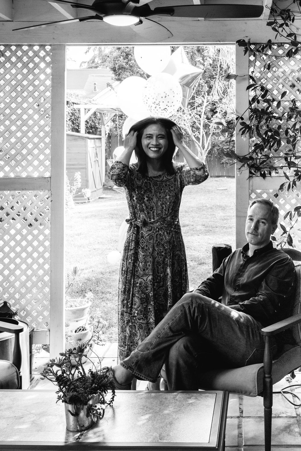 Black and white photo of two people relaxing on a covered porch, one standing in a patterned dress with a hat.