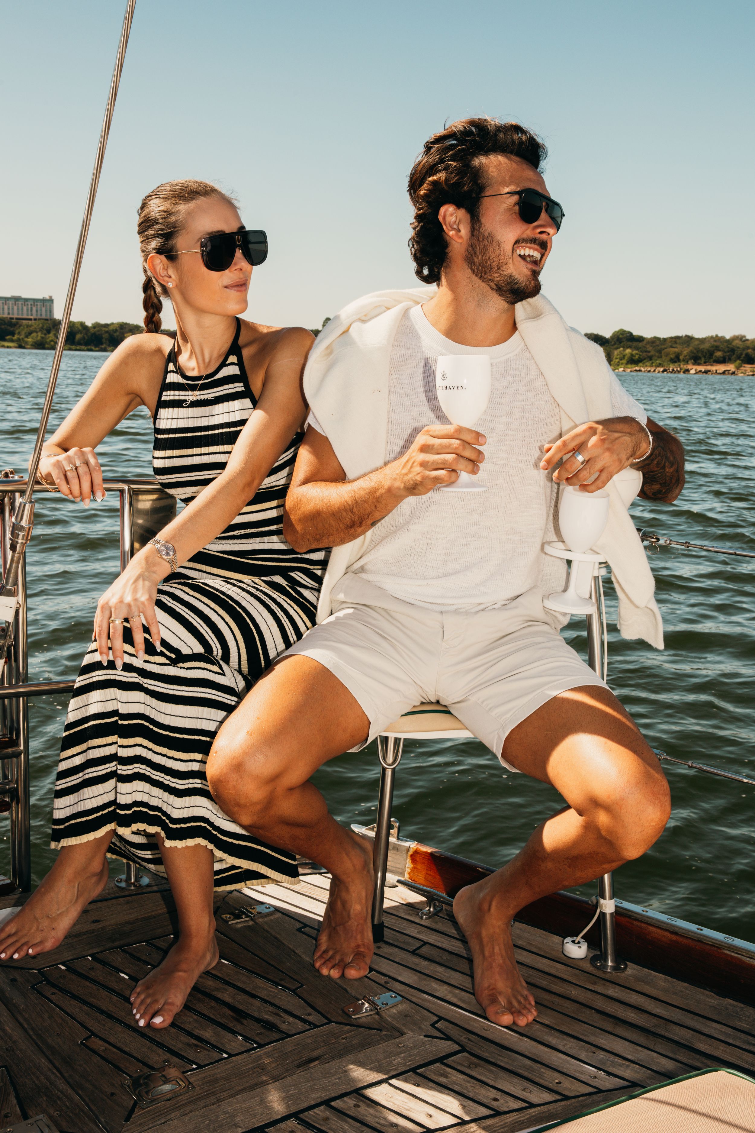 Two people enjoying drinks while relaxing on a sailboat deck during a sunny summer day.