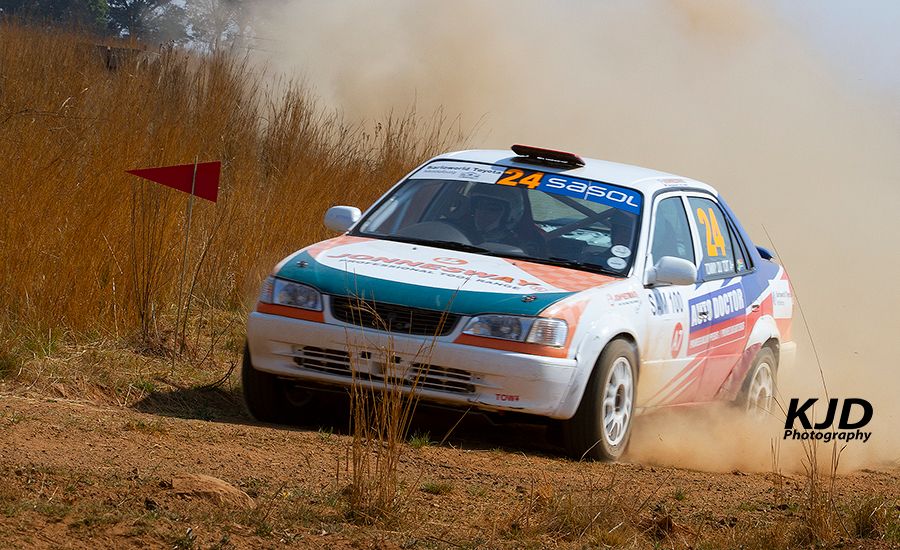 Rally car racing through a dusty dirt track with tall brown grass in the background.