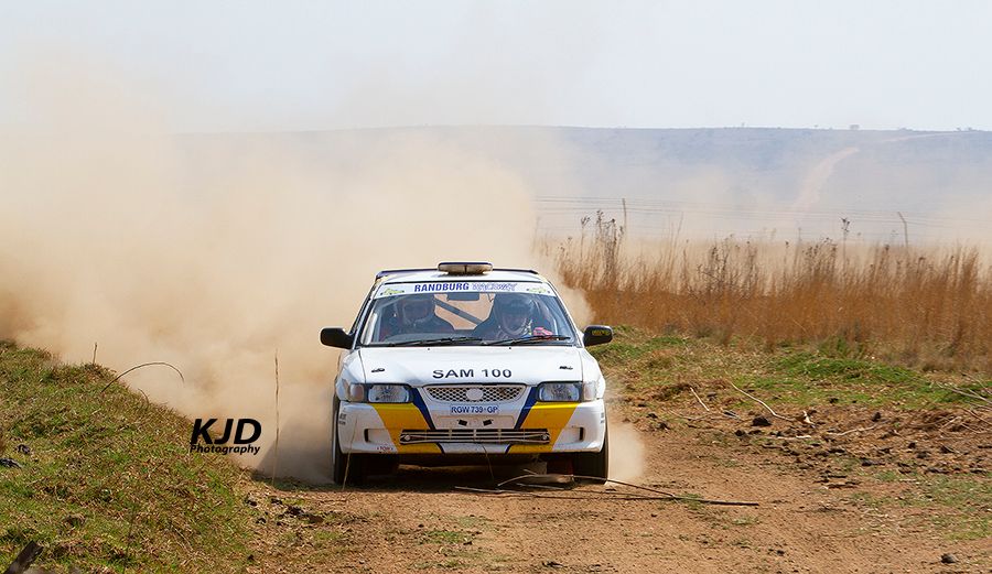 A rally car races on a dusty dirt track, kicking up clouds of dust while speeding through a rural landscape.