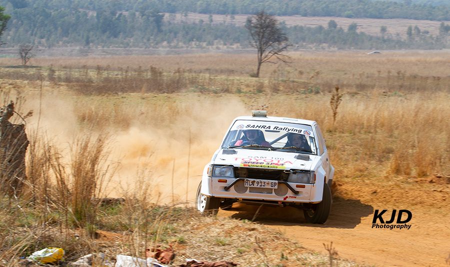 A white rally car kicks up dust while racing through a dirt track in a dry grassland terrain.