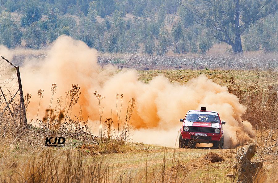 Rally car kicks up orange dust while racing through a dry, rural course surrounded by wild brush and trees.