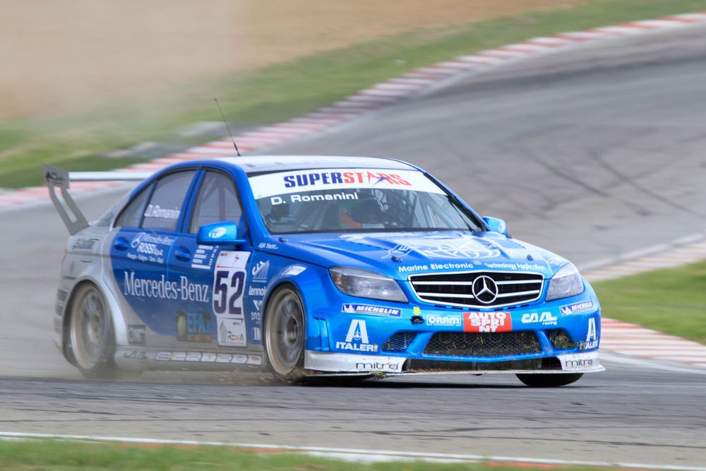 A blue Mercedes-Benz C-Class race car drifts through a corner on a race track with tire smoke trailing behind.
