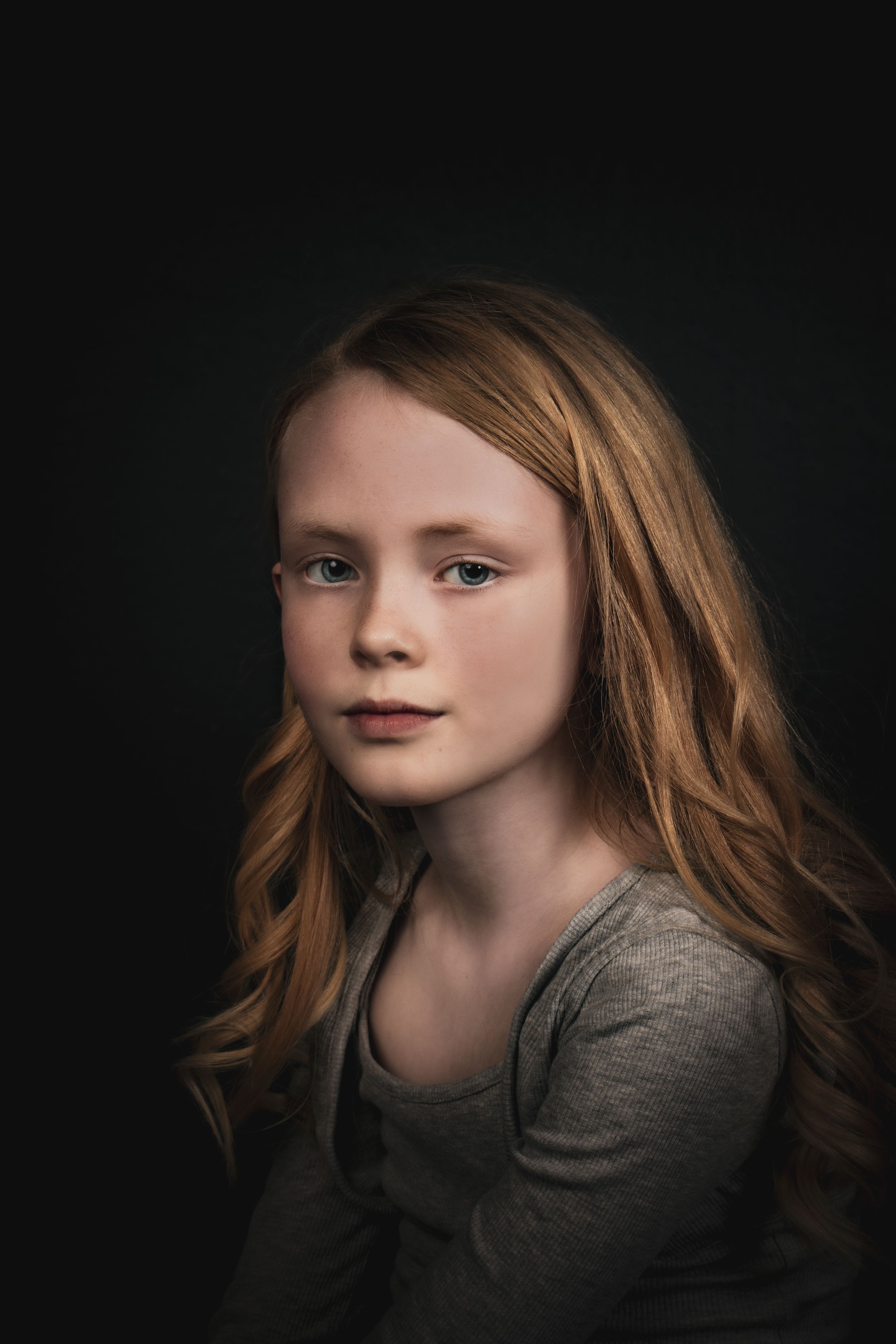 Portrait against dark background shows young person with flowing red hair wearing grey shirt in dramatic lighting.