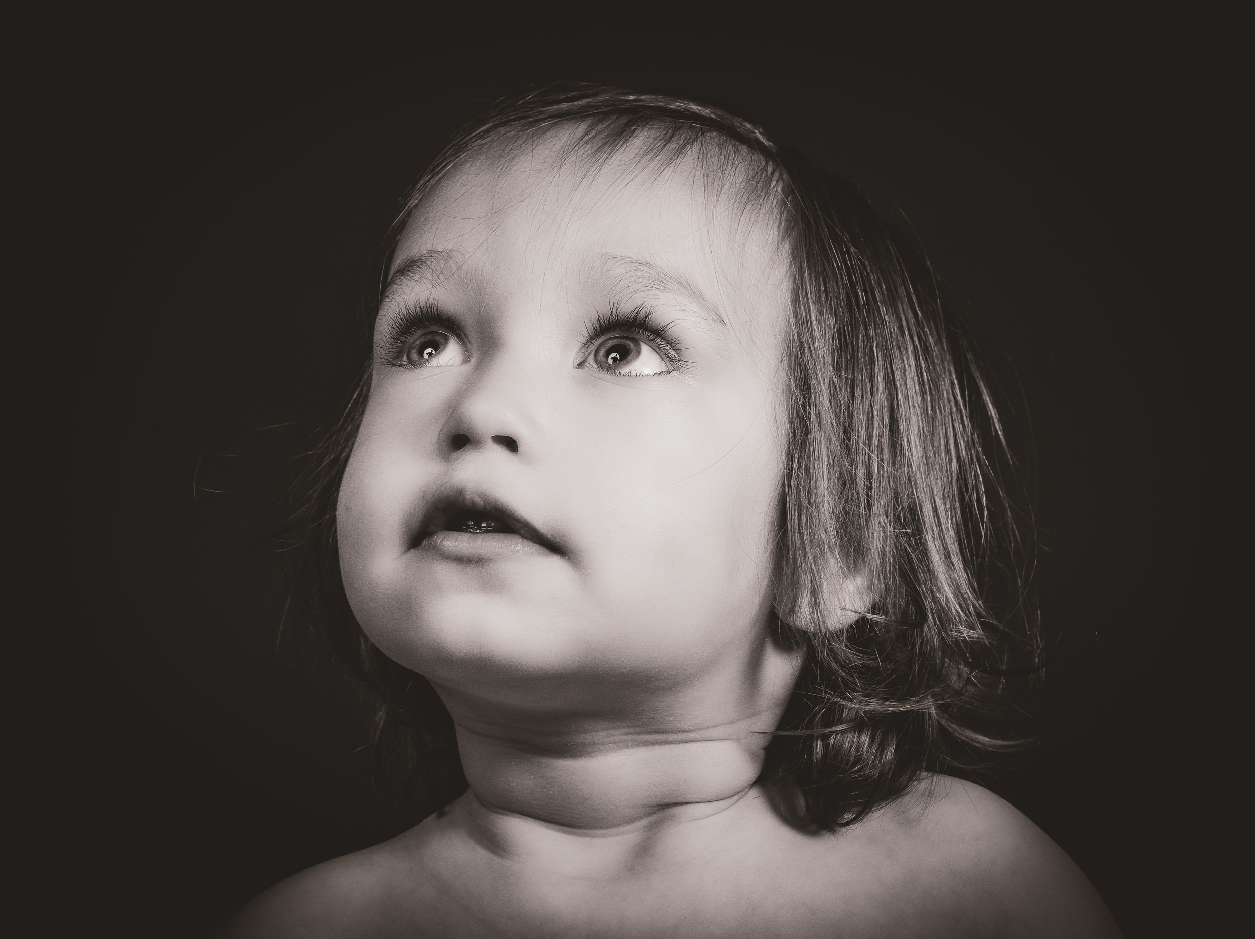 Fine art black and white portrait of young child looking up with wonder, captured in dramatic lighting.