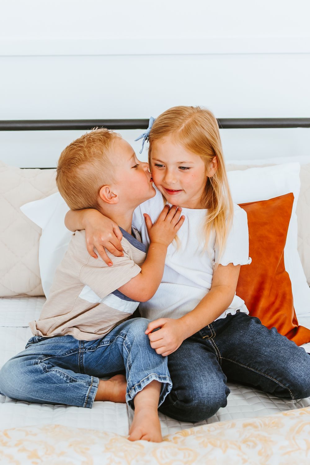 Two young children share a sweet moment on a couch with orange and white pillows.