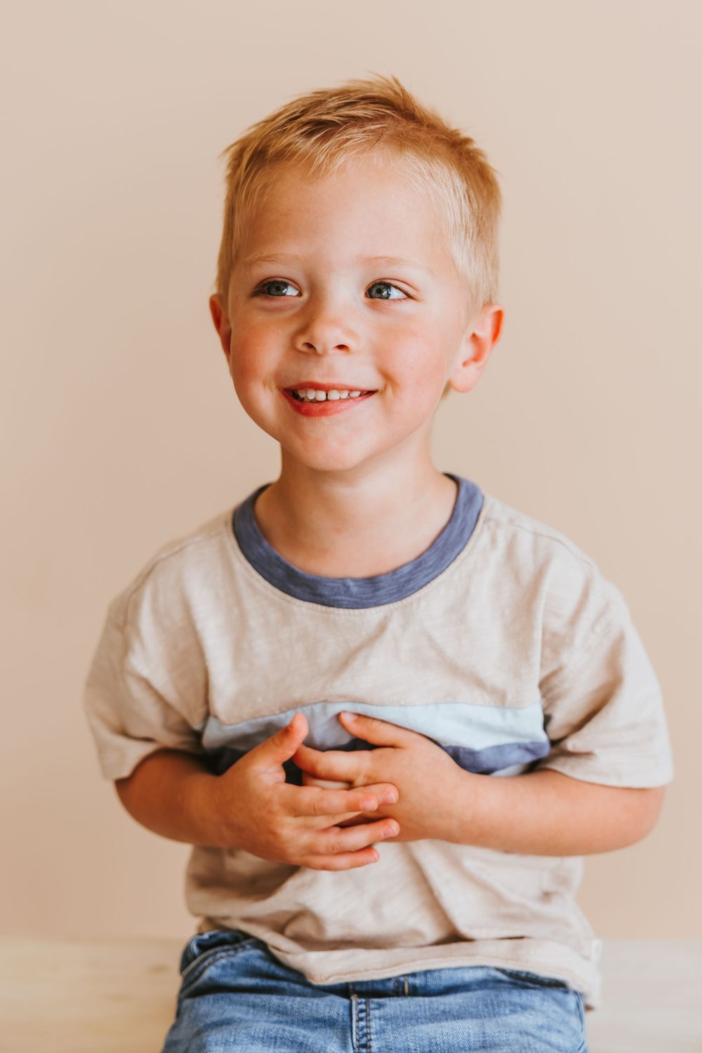 A young child wearing a gray t-shirt and jeans smiles brightly while clasping their hands together.