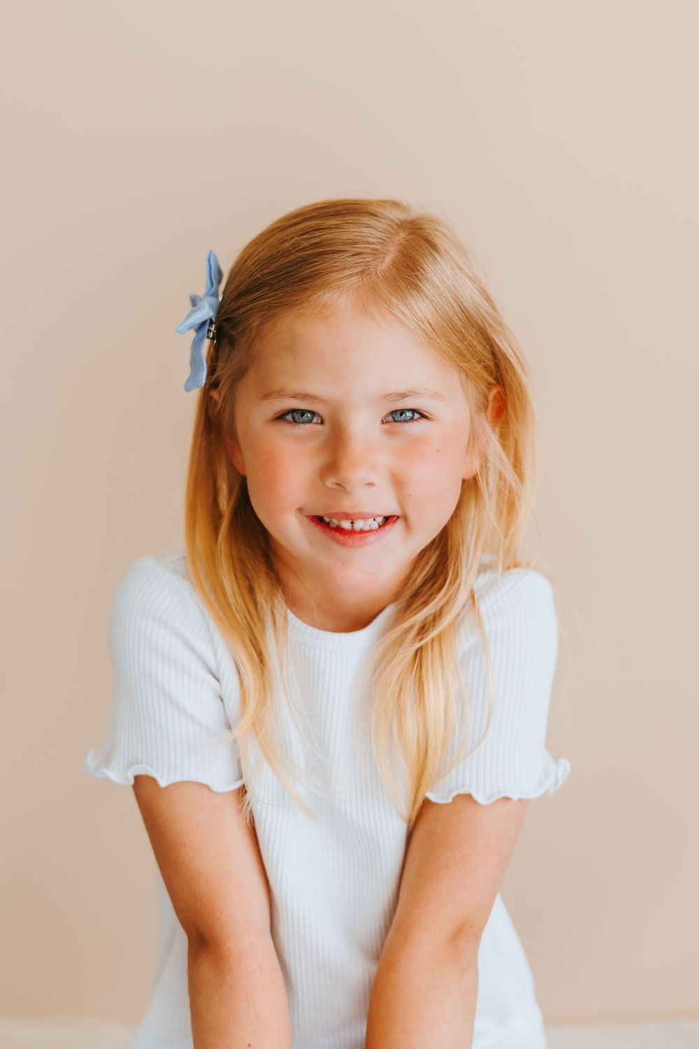 A cheerful child in a white ruffled shirt and blue hair clip beams at the camera against a neutral background.