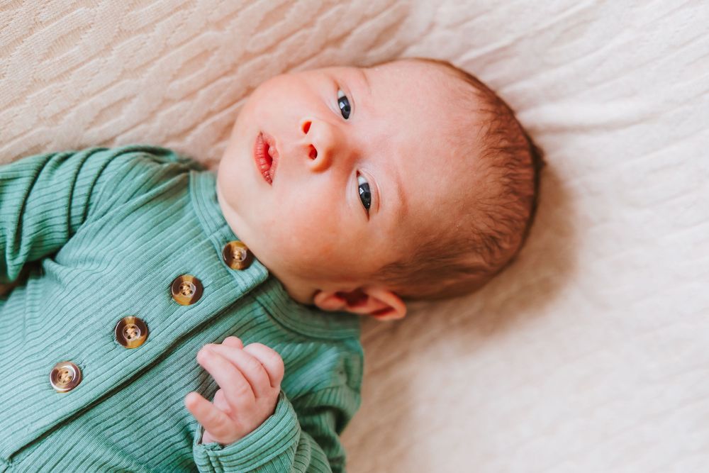 Child in a green knit cardigan rests on a white bedding while looking towards the camera.