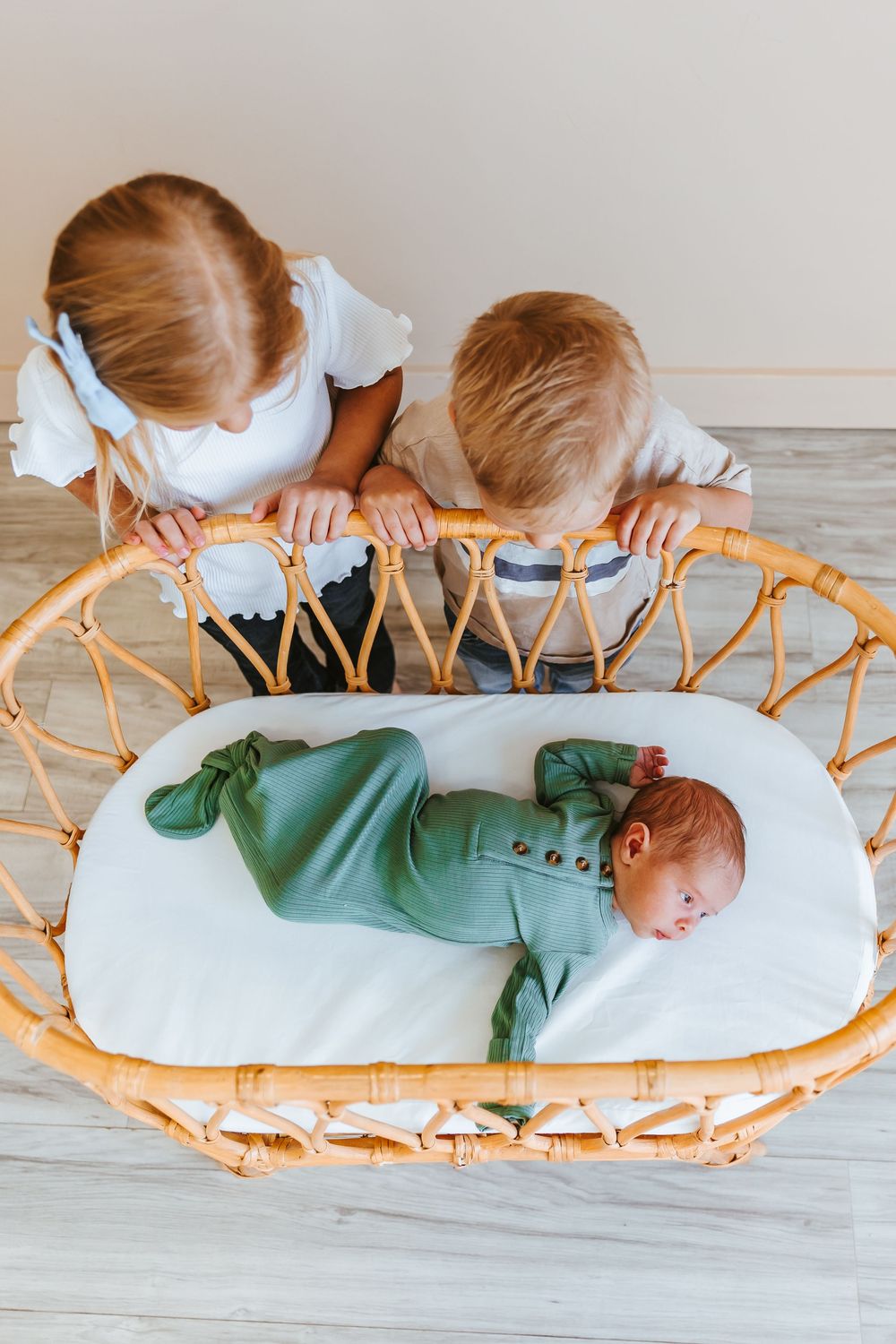 Young children looking down at baby on a woven rattan lounger with white cushions.