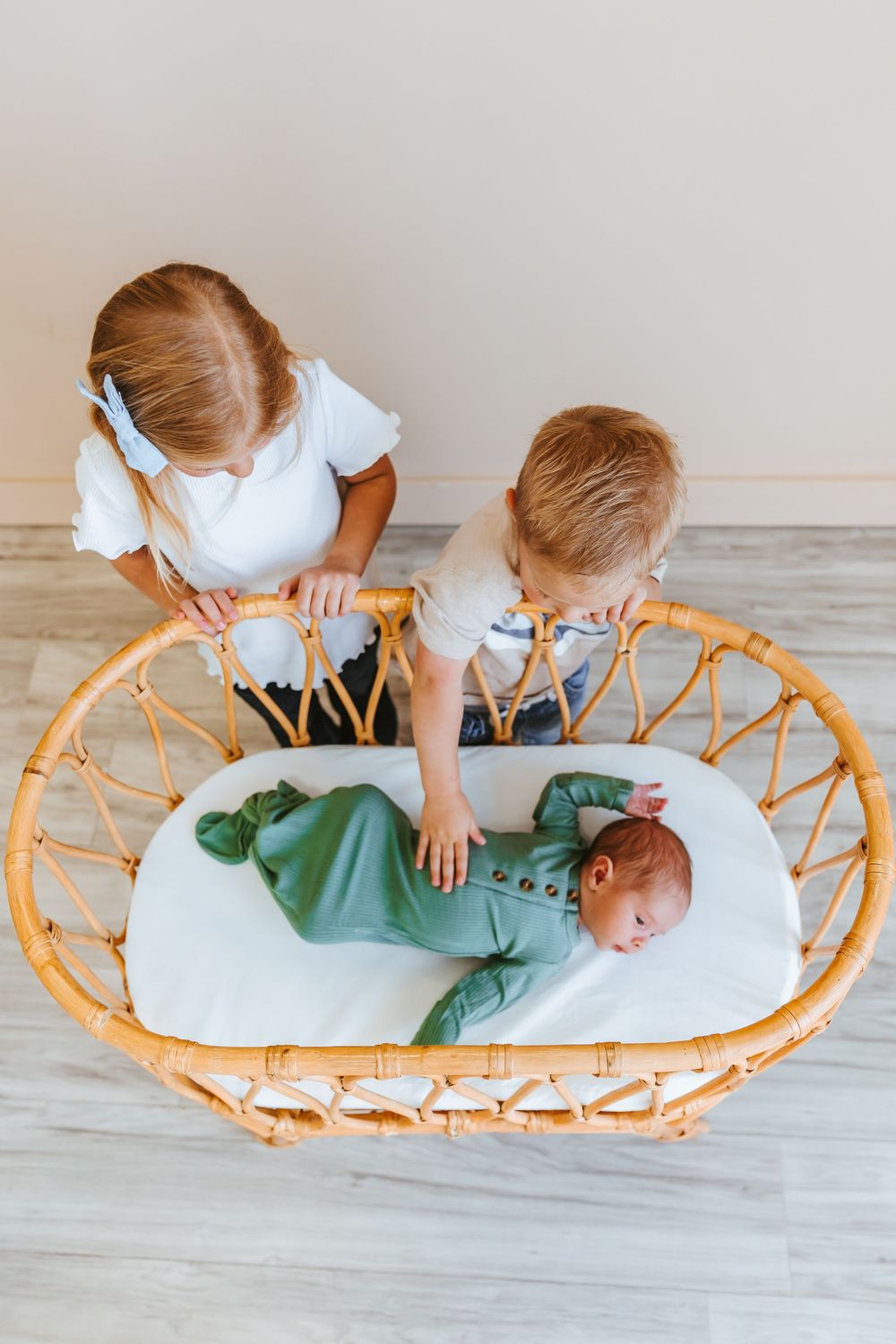 Siblings interacting with baby lying on round rattan lounger with white cushion.