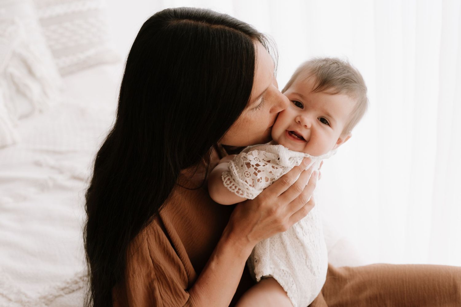 Woman in brown top lovingly kisses smiling baby dressed in white against light background.