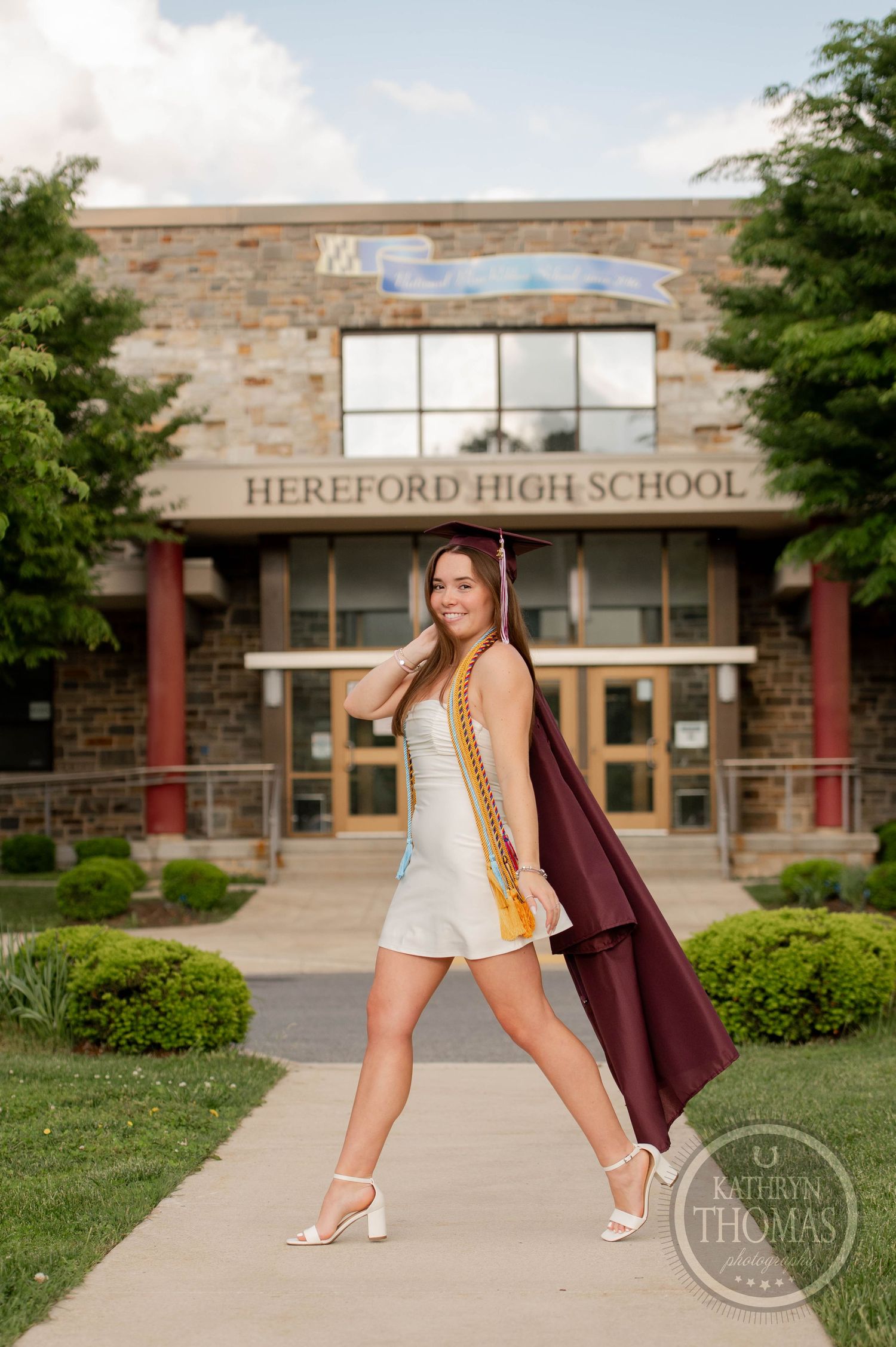 A high school cap and gown graduation portrait session featuring white high heels against the brick exterior of Hereford High School.