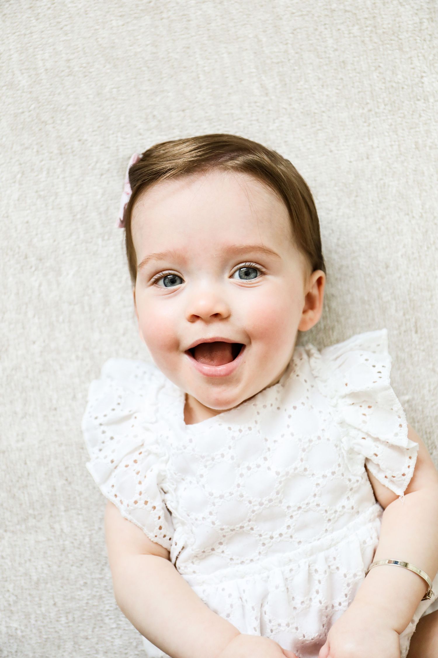 A happy baby wearing a white eyelet lace dress smiles brightly against a light coloured background, during birthday shoot in Nottingham
