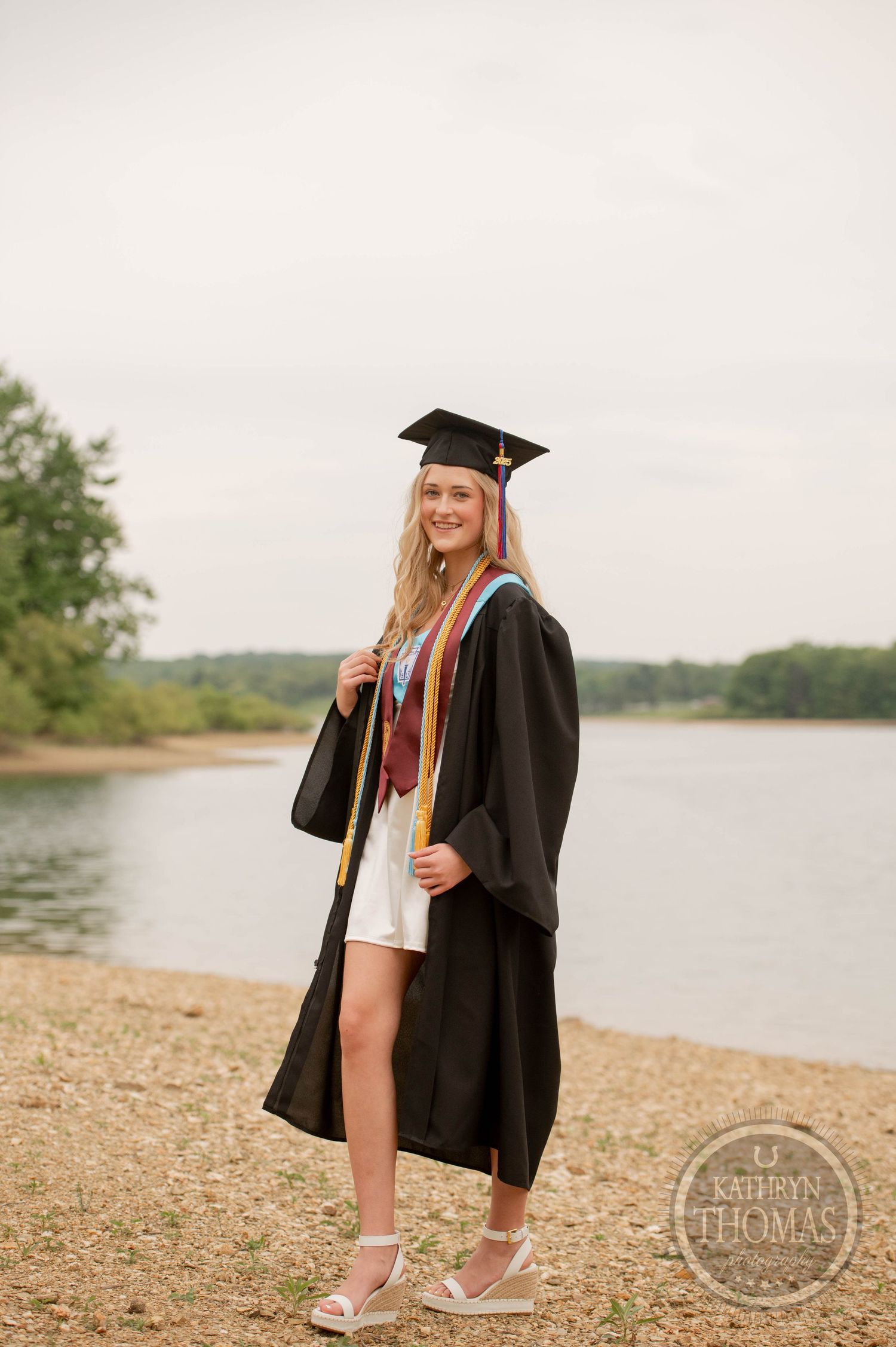 Graduate wearing cap and gown poses on beach shoreline near lake with trees in background.