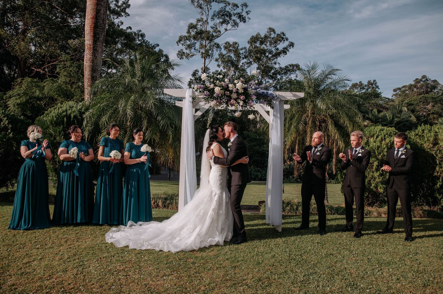 Wedding ceremony at elegant outdoor garden venue with bridal party in teal dresses and dark suits under white pergola.