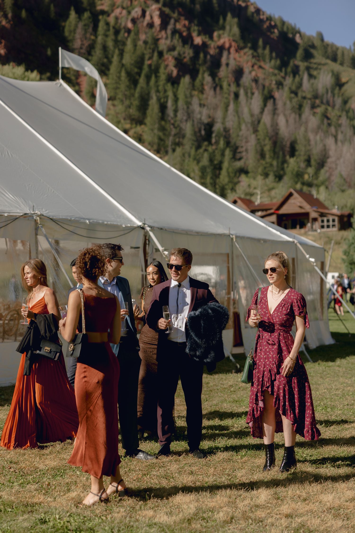 A group of wedding guests gather outside a white tent at an outdoor mountain venue during a sunny afternoon.