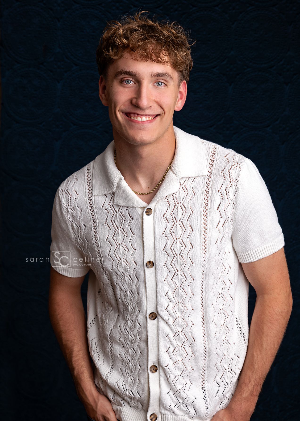 Professional studio portrait of someone in a white knit polo shirt with decorative pattern against dark background.