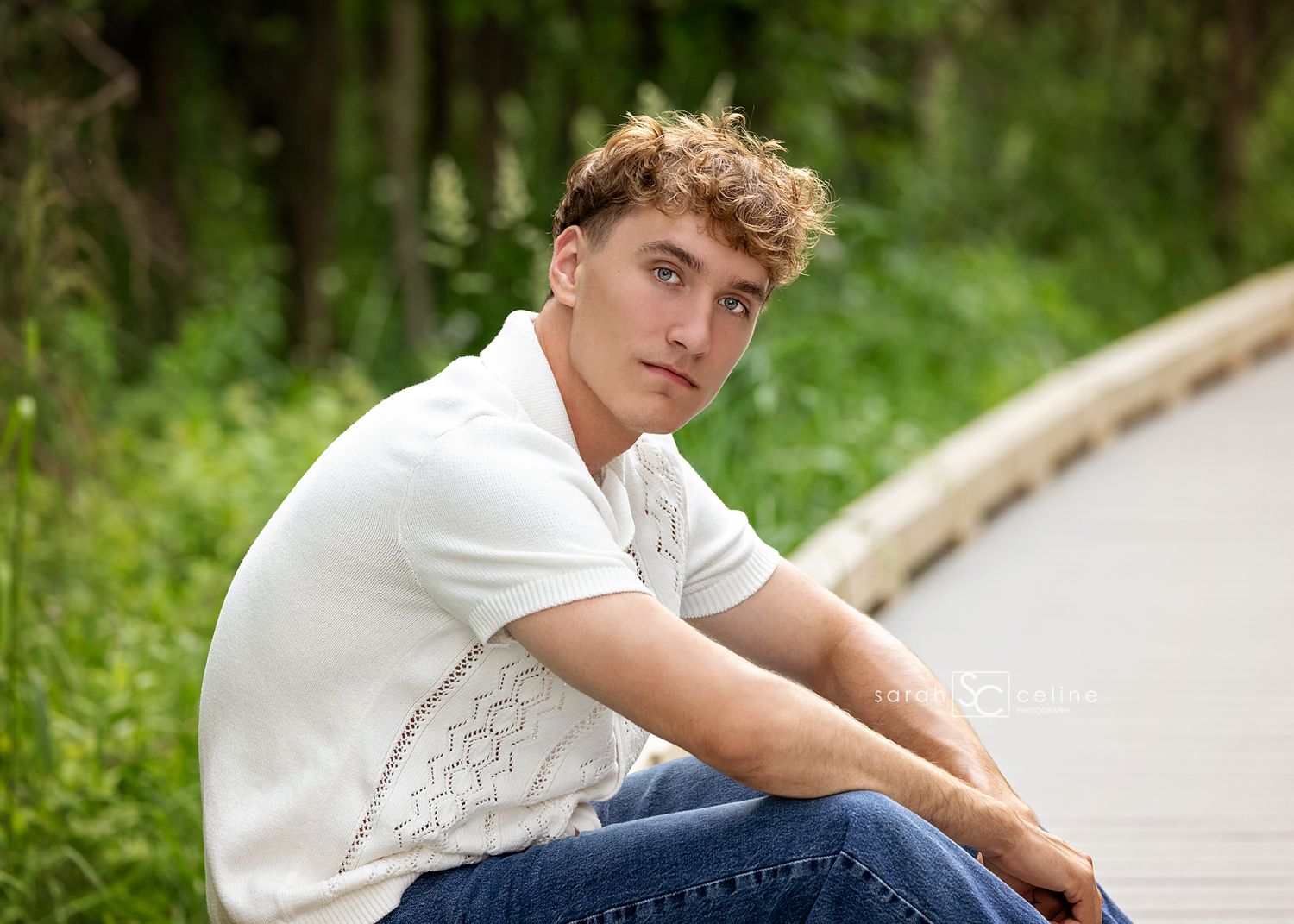 Young person in white t-shirt sitting casually outdoors against natural green background.
