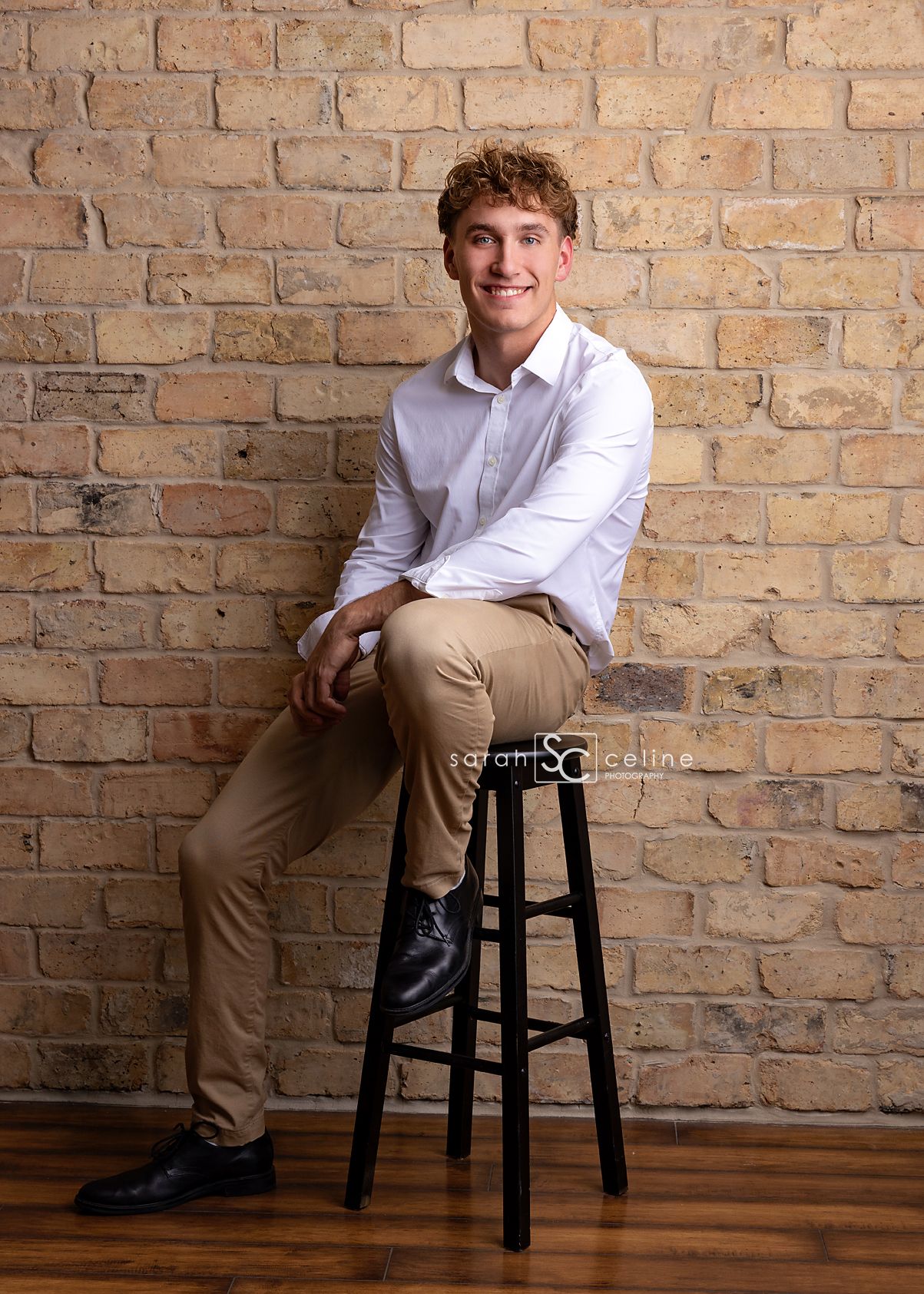 A person in a white button-down shirt and khaki pants sits casually on a dark wooden stool against a brick wall background.