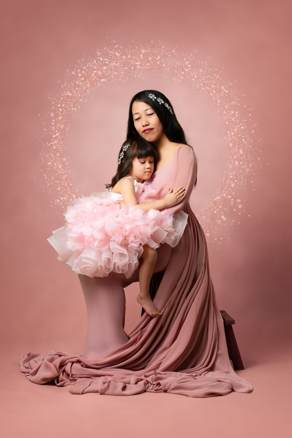 Mother in pink gown tenderly holds young daughter in fluffy pink dress against a soft pink backdrop with sparkles.
