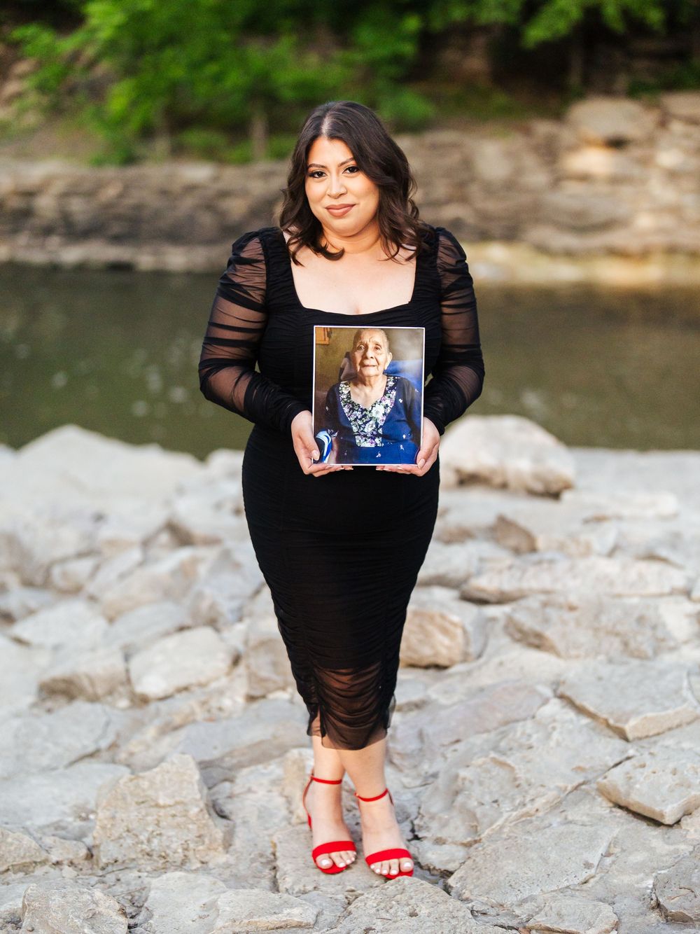 Person in black dress stands on rocky riverside holding a photo frame with red shoes visible.