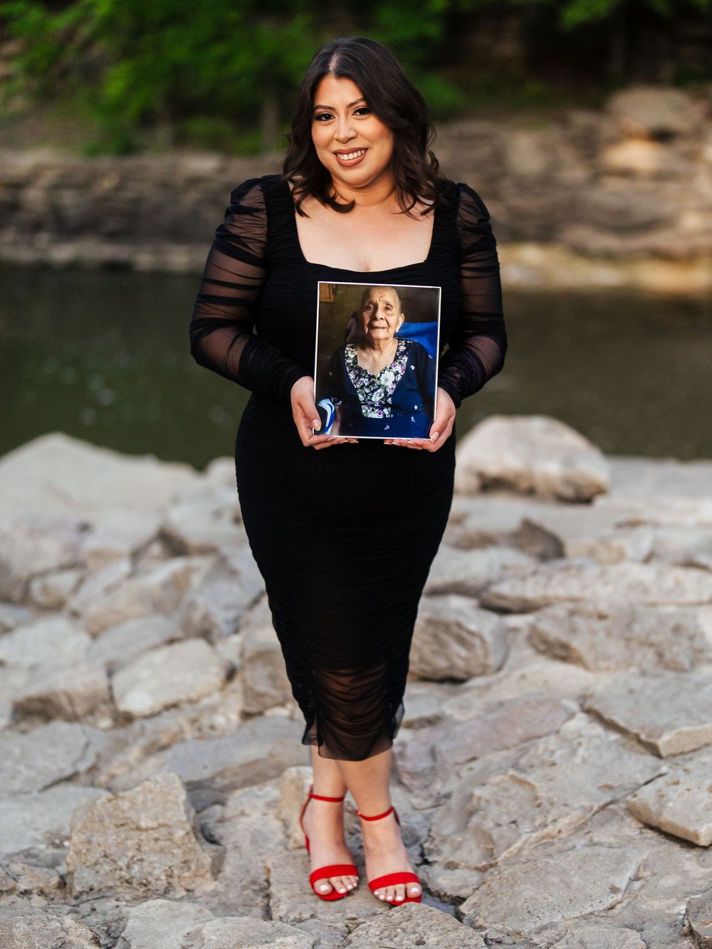 A person in a black lace dress holds a framed photograph while standing on rocky terrain near water.