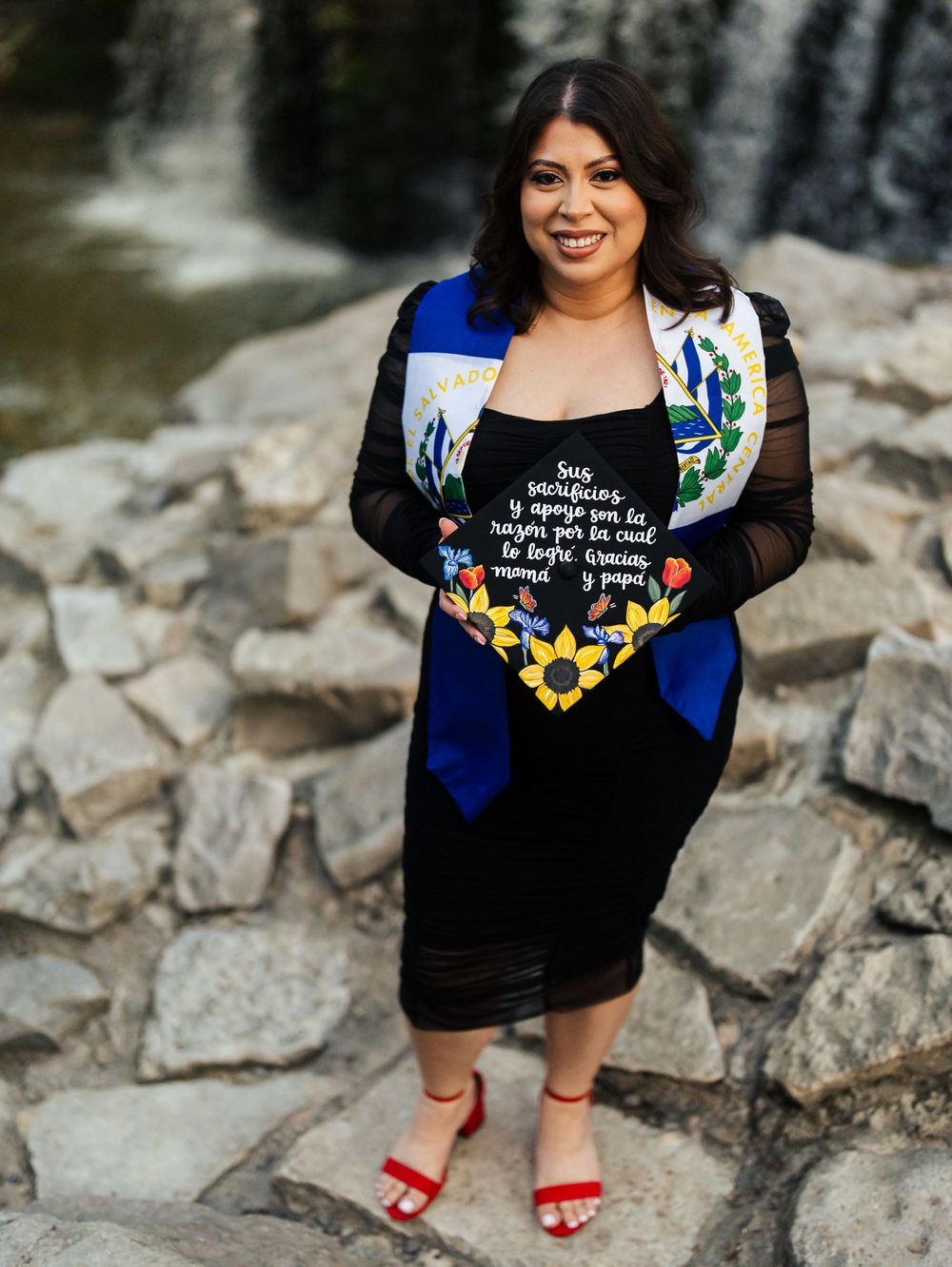 A person in a black floral dress with red heels poses outdoors by a rock wall and waterfall.