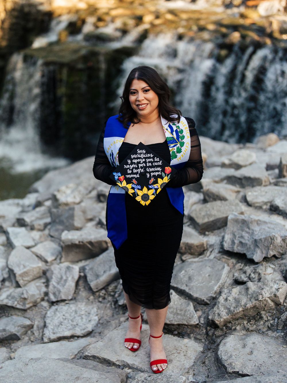 A graduate in a blue and black dress poses in front of a cascading waterfall on rocky terrain.
