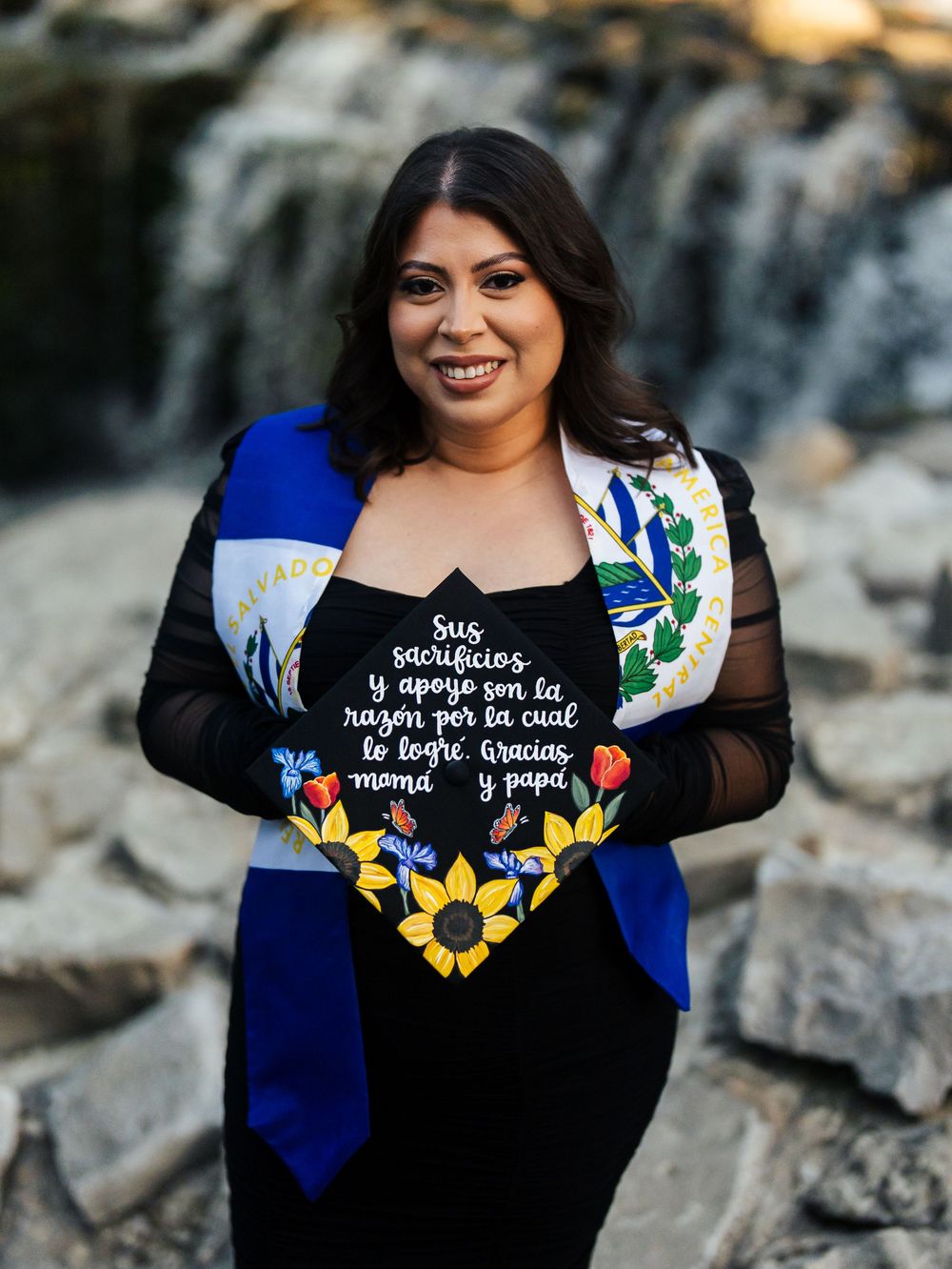 Graduate wearing decorated black graduation cap and gown with floral sunflower design against rocky outdoor background.