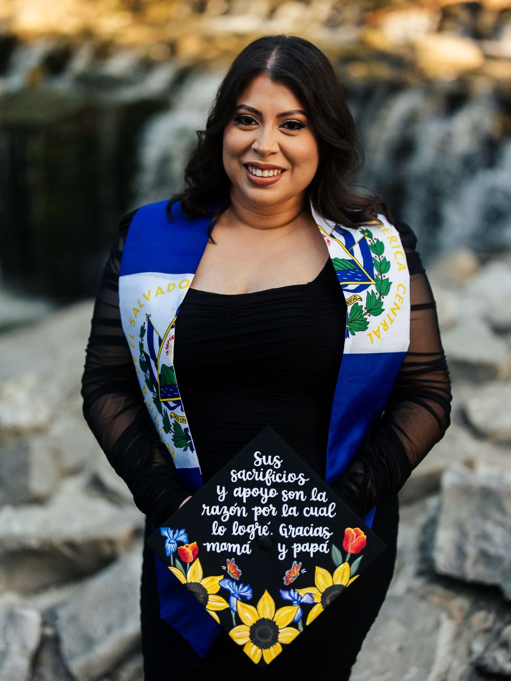 Woman in black outfit with embroidered flowers and Spanish text posing outdoors against a natural backdrop.