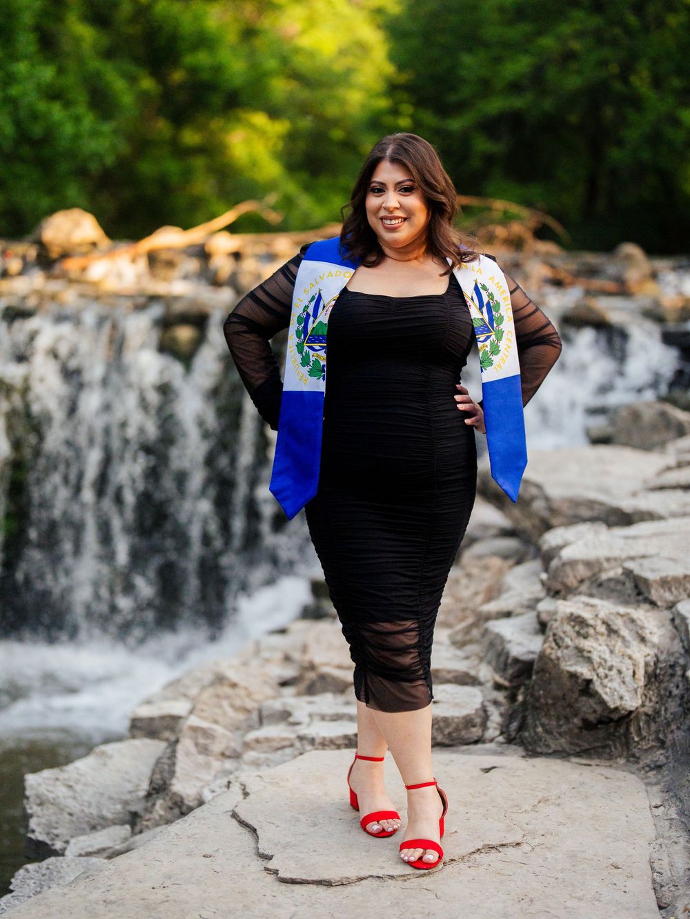 A figure in a black dress and blue jacket stands confidently on rocks near a waterfall wearing bright red heels.