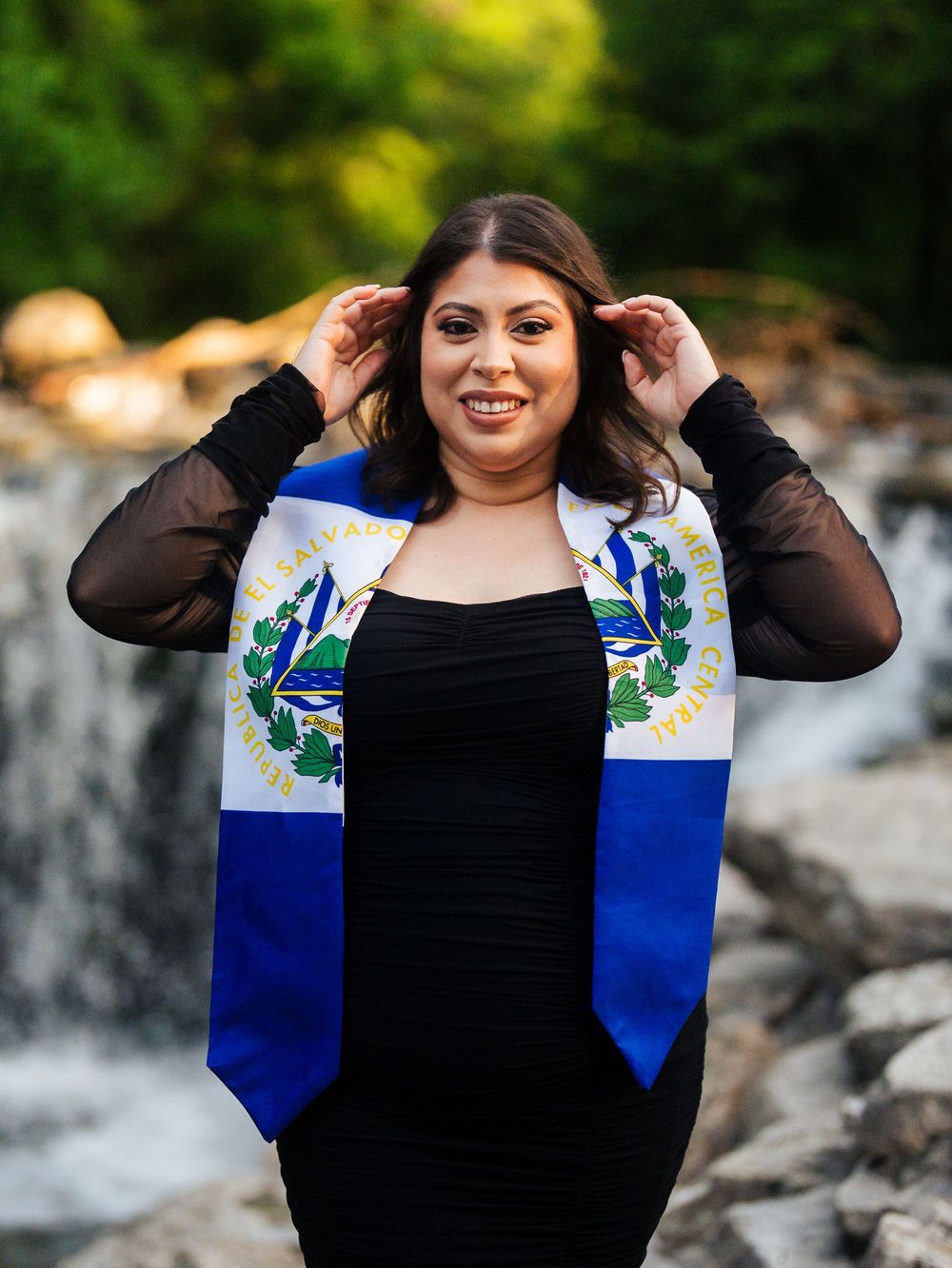 A person in a black dress and blue floral cardigan stands in front of a waterfall on a sunny day.