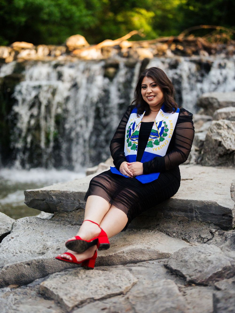 Graduate in black dress and blue stole sits on rocks by cascading waterfall during outdoor photo shoot.