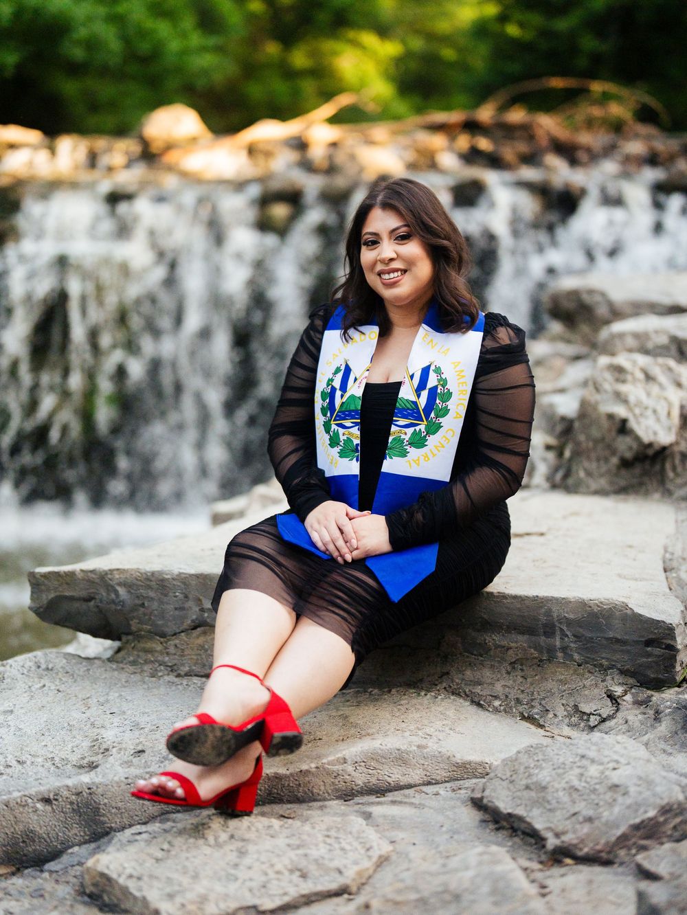 Graduate sits on rocky ledge wearing blue graduation stole and red shoes near cascading waterfall backdrop.