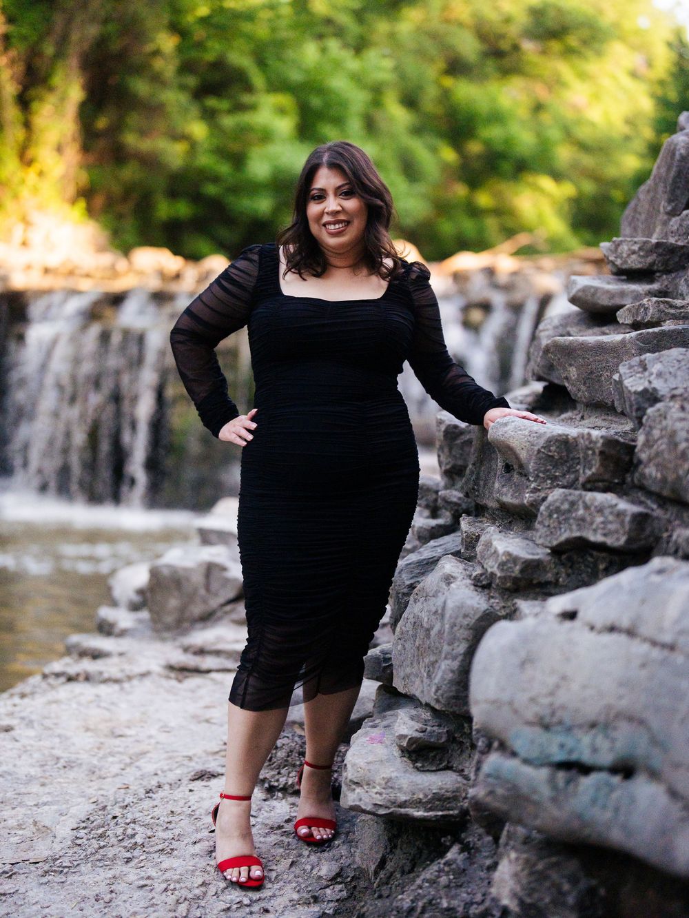 Person in a black dress poses against rock formations near a waterfall while wearing red sandals.