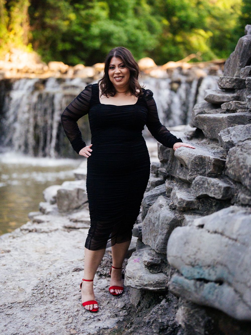 Person wearing a black top sits on limestone rocks by a flowing stream on a sunny summer day, showing red shoes.