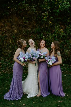Bridesmaids in purple dresses hold blue and purple bouquets while sitting on grass.