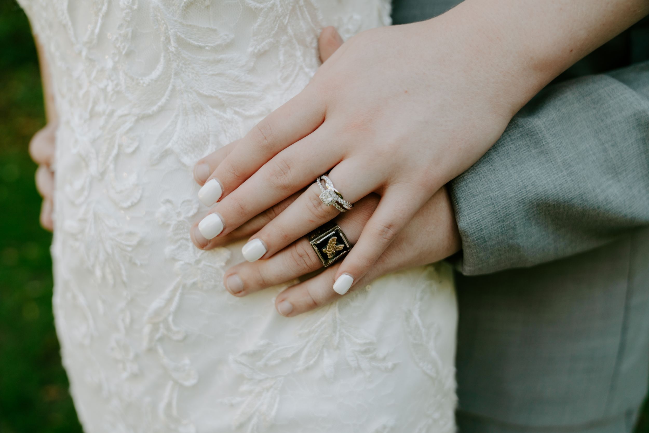 Close-up of hands showing wedding rings against white lace dress and gray suit.