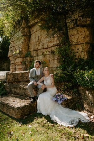 Wedding couple poses against stone wall backdrop in garden setting with purple flowers.