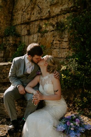 Intimate wedding moment captured against weathered stone wall with purple floral bouquet.