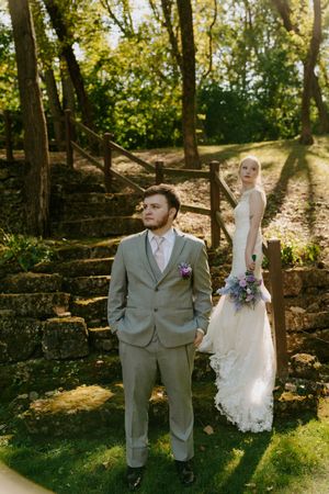 Wedding couple poses in a wooded area with stone walls and dappled sunlight filtering through trees.