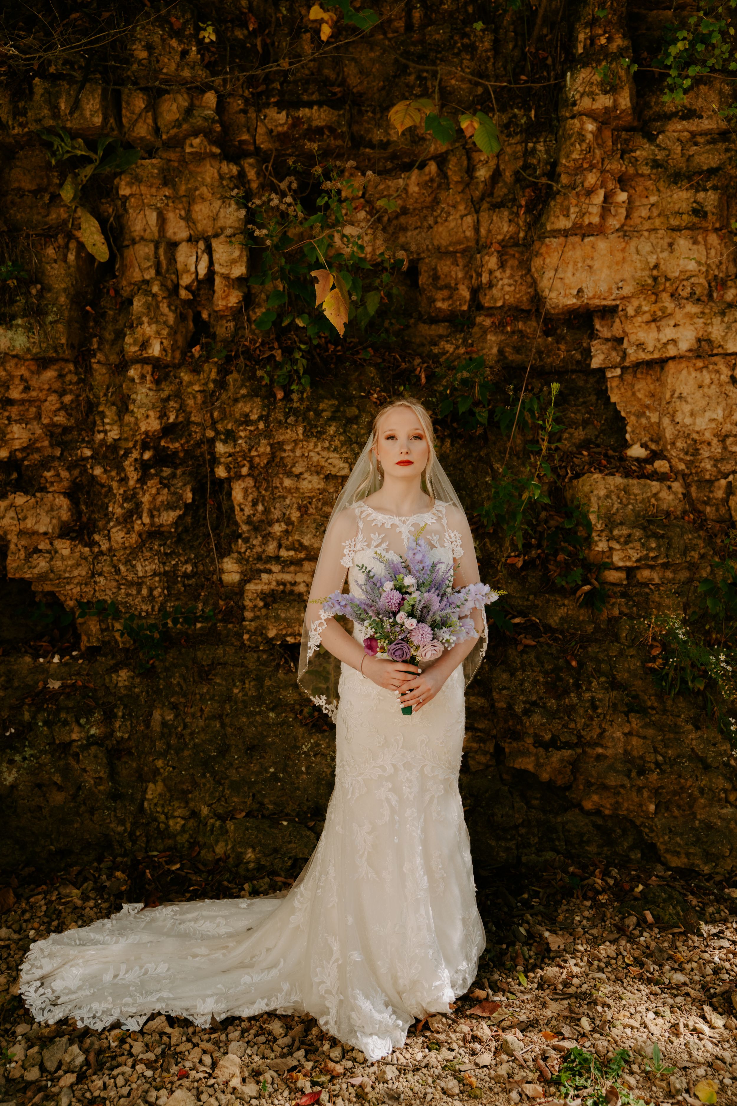 Bride in lace wedding gown holding purple bouquet against stone wall backdrop.